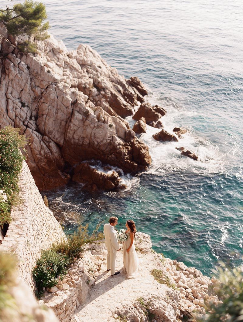 Bride and groom embracing on a sunlit Amalfi Coast cliffside overlooking turquoise Mediterranean waters during their luxury destination wedding